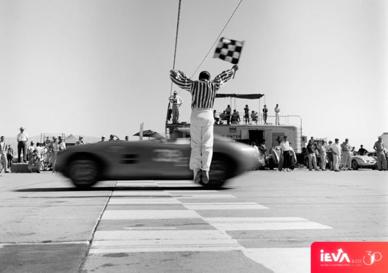 Black-and-white photograph of a race official waving a checkered flag as a car crosses the finish line, with an IEVA & Co 30th-anniversary mark in the lower corner.